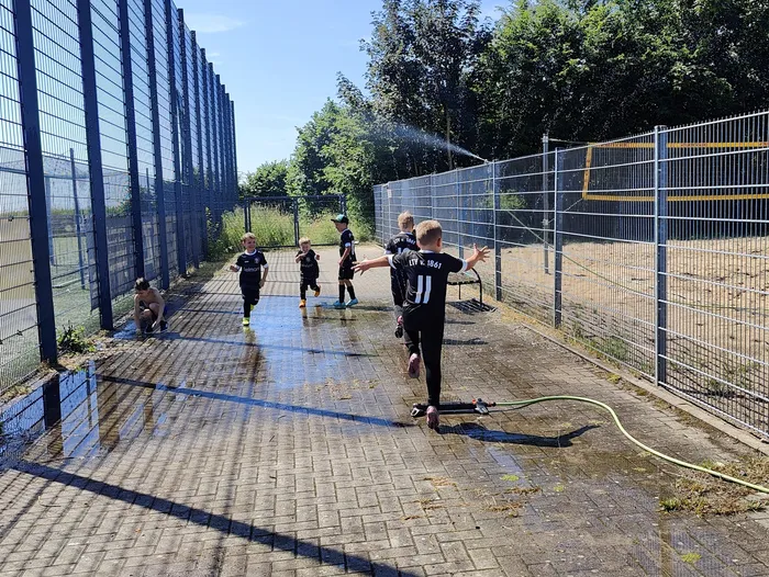 Fünf junge Fußballspieler in schwarzen Trikots und Shorts laufen auf einem nassen, gepflasterten Platz. Ein Spieler rennt im Vordergrund auf eine Wasserfontäne zu, während die anderen hinter ihm jubeln. Im Hintergrund eine hohe blaue Metallwand und Bäume.