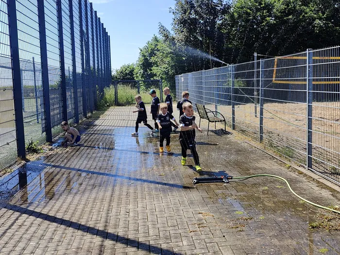 Junge Fußballspieler in schwarzen Trikots laufen auf einem nassen, gepflasterten Platz, während ein Sprinkler sie mit Wasser besprüht. Im Hintergrund ein Zaun und Büsche.
