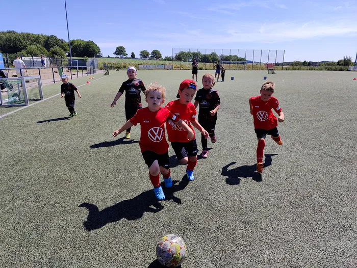 Eine Gruppe junger Fußballspieler in roten Trikots und schwarzen Shorts trainiert auf einem grünen Kunstrasenplatz. Im Vordergrund dribbelt ein Spieler den Ball. Im Hintergrund sind weitere Spieler und Trainingshütchen zu sehen.
