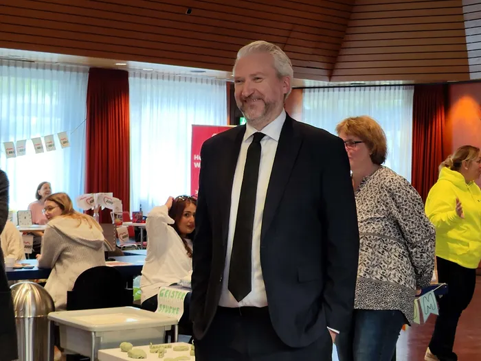 Ein lächelnder Mann im dunklen Anzug steht in einem Raum mit roten Vorhängen, im Hintergrund sind weitere Personen an Tischen zu sehen.