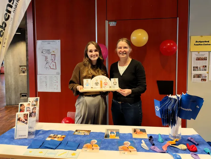 Zwei Frauen präsentieren Informationsmaterialien und blaue Taschen an einem Stand beim Neugeborenen-Empfang.