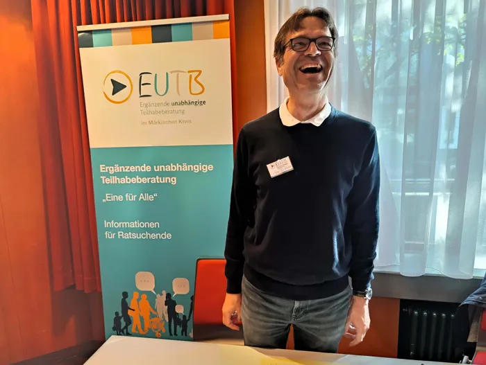 Ein lachender Mann mit Brille steht vor einem Banner der EUTB (Ergänzende unabhängige Teilhabeberatung).