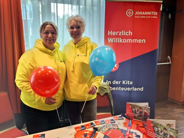 Zwei Frauen in gelben Kapuzenpullovern halten einen roten und einen blauen Ballon vor einem roten Johanniter-Willkommensbanner.