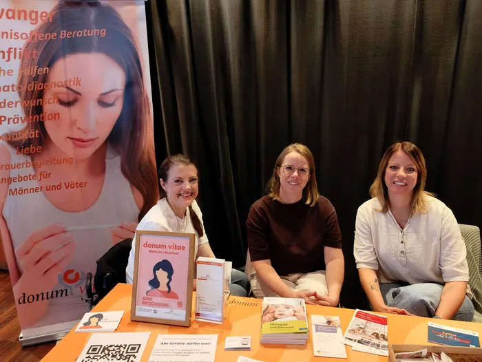 Drei lächelnde Frauen sitzen an einem Tisch mit Informationsmaterialien von „donum vitae“ vor einem großen Banner.