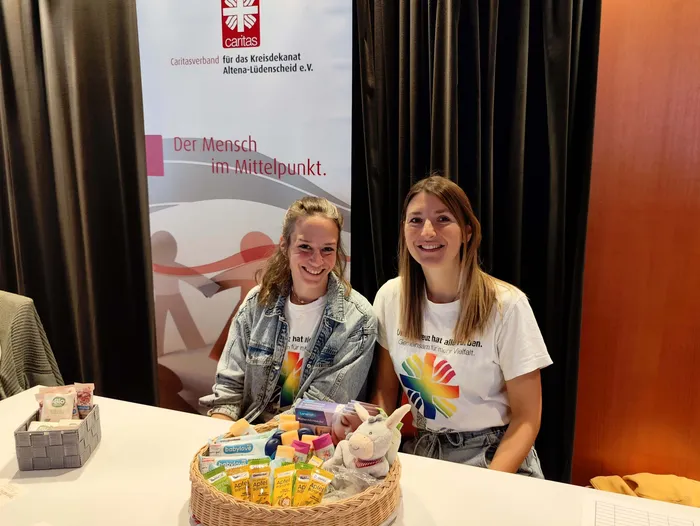 Zwei lächelnde Frauen sitzen hinter einem Tisch mit einem Korb voller Babyprodukte vor einem Caritas-Banner.