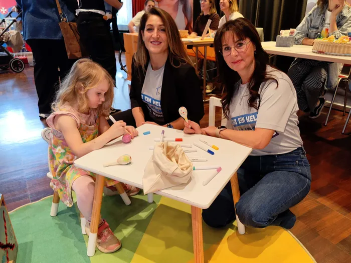 Ein kleines Mädchen und zwei Frauen sitzen an einem Tisch und spielen mit Maracas und Stöcken.