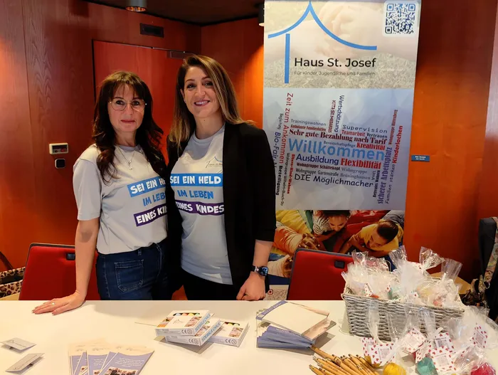 Zwei Frauen stehen hinter einem Tisch mit Flyern und Geschenken vor einem Banner von Haus St. Josef. Ihre T-Shirts tragen die Aufschrift „Sei ein Held im Leben eines Kindes“.