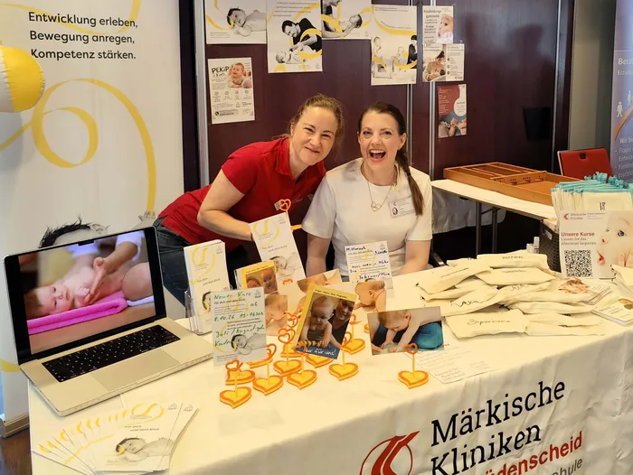 Zwei lächelnde Frauen stehen hinter einem Infostand der Märkischen Kliniken Lüdenscheid mit Broschüren und einem Laptop.