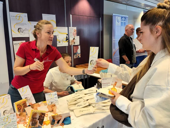 Eine Frau in einem roten Poloshirt überreicht einer Besucherin einen Flyer an einem Infostand beim Neugeborenen-Empfang.