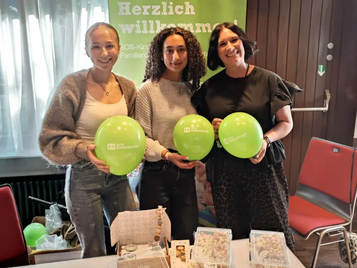 Drei lächelnde Frauen halten grüne Luftballons mit der Aufschrift „SOS Kinderdorf“.