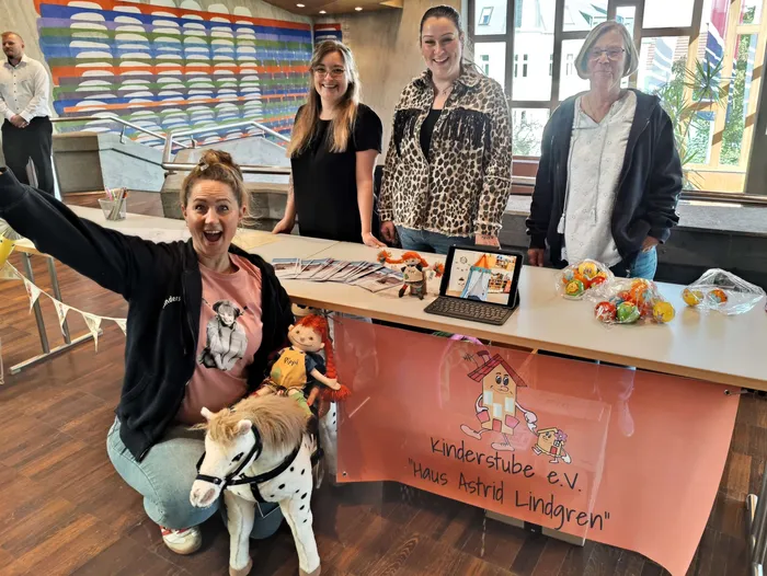 Vier Frauen posieren hinter einem Tisch mit einem Banner der „Kinderstube e.V. Haus Astrid Lindgren“, auf dem Puppen und Süßigkeiten liegen.