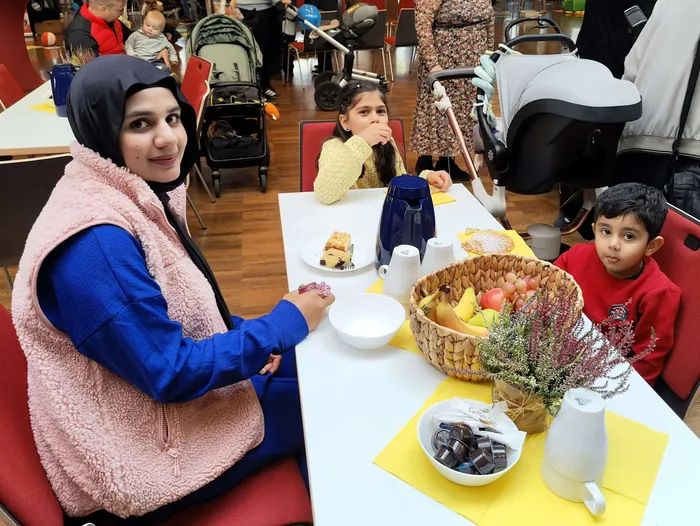 Eine Frau mit Hijab und rosa Weste sitzt an einem Tisch mit Speisen und Getränken bei einem Neugeborenen-Empfang.