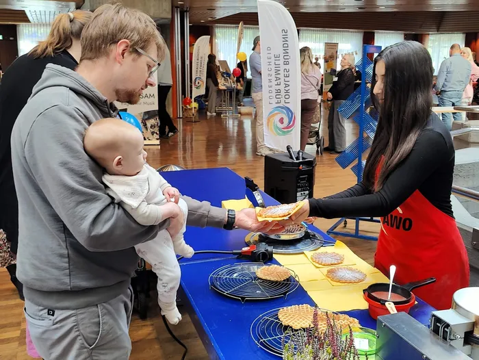 Ein Mann hält ein Baby und nimmt eine Waffel von einer Frau in einer roten Schürze an einem Stand entgegen.