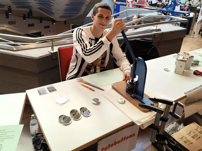 Ein junger Mann bedient eine blaue Button-Presse an einem Tisch mit Bastelmaterialien und fertigen Buttons.