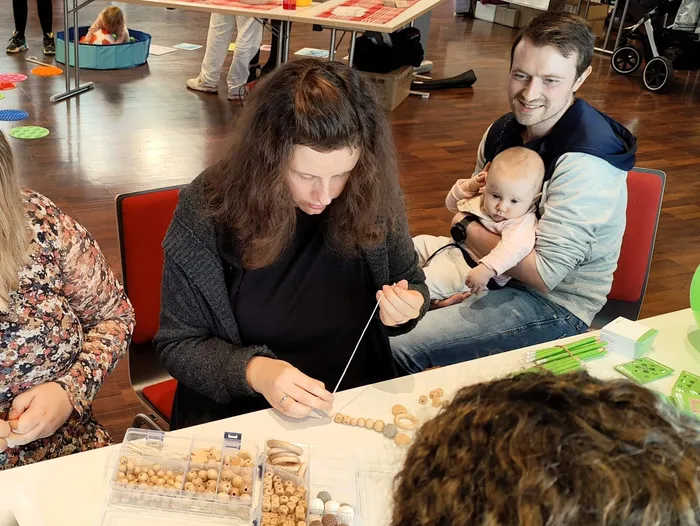 Eine Frau arbeitet mit kleinen Holzperlen an einem Tisch, während ein Mann ein Baby im Arm hält.