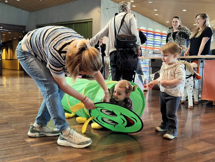Eine Frau hilft einem Kind beim Krabbeln durch einen grünen Spieltunnel bei einem Neugeborenen-Empfang.