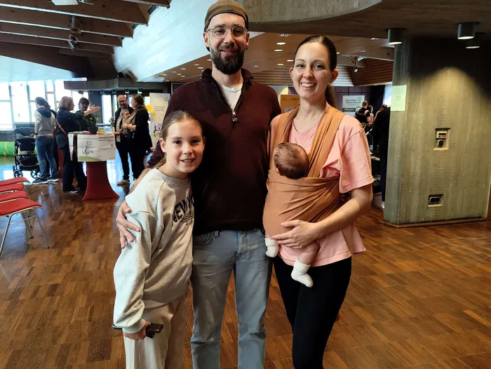 Eine lächelnde Familie mit einem Baby in einer Trage posiert bei einem Neugeborenen-Empfang in einem Kulturhaus.
