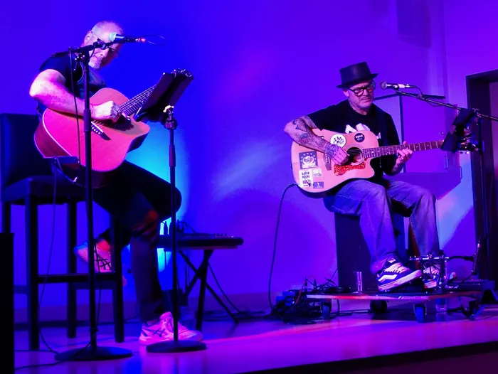 Tobias Janssen und Jiri „George“ Schneider spielen Akustikgitarren auf einer Bühne unter blauem und violettem Licht.
