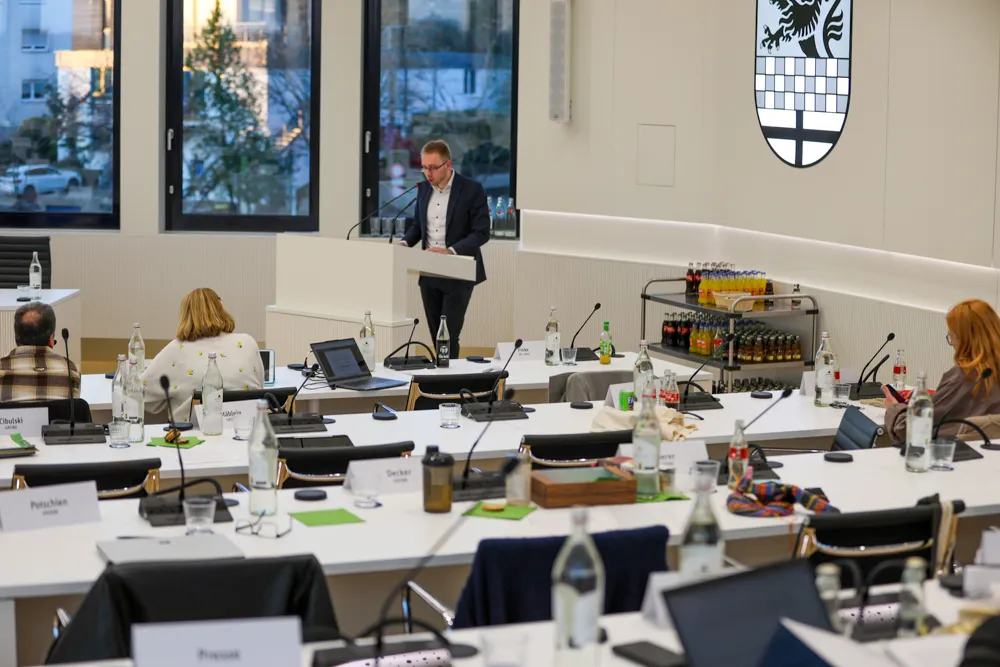 Mann am Rednerpult vor Reihen von Sitzplätzen, einige davon leer, während andere von Personen besetzt sind. Im Hintergrund ein Wappen an der Wand.