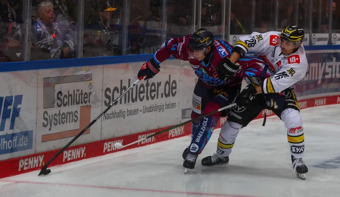 Ein Spieler der Iserlohn Roosters in Rot-Blau und ein Spieler der Krefeld Pinguine in Weiß im Zweikampf auf dem Eis.