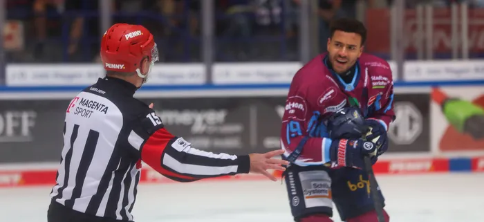 Ein Eishockey-Schiedsrichter gestikuliert gegenüber einem Spieler der Iserlohn Roosters in einem weinroten Trikot auf dem Eis.