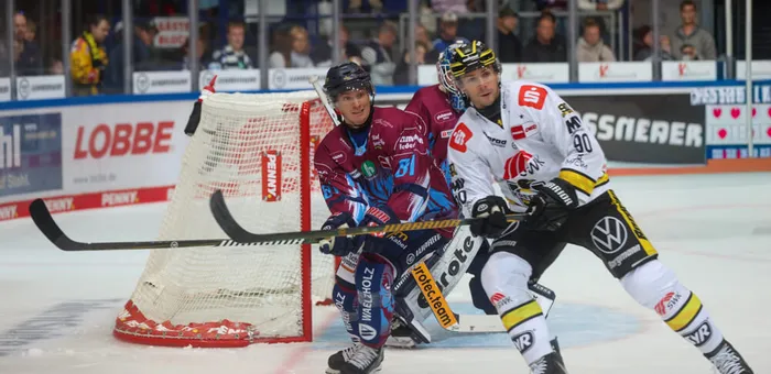Ein Eishockeyspieler in weißem Trikot und ein Torwart in weinroter Kleidung im Zweikampf vor dem Tor.