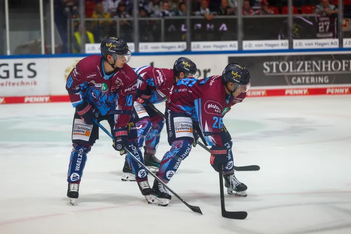 Drei Eishockeyspieler der Iserlohn Roosters in blau-roten Trikots in Spielaktion auf dem Eis.