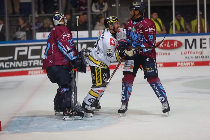 Eishockeyspieler in weinroten und weißen Trikots stehen in einer Spielsituation vor dem Tor auf dem Eis.
