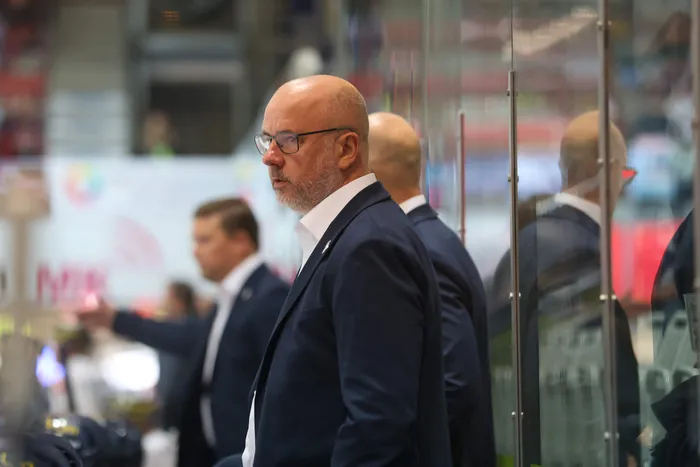 Ein glatzköpfiger Mann mit Brille und Bart im dunklen Anzug blickt in einer Eishalle zur Seite.