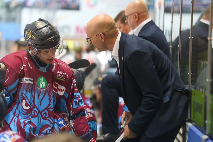 Ein Eishockeyspieler der Iserlohn Roosters in weinrotem Trikot wird von einem Mann im dunklen Anzug angesprochen.