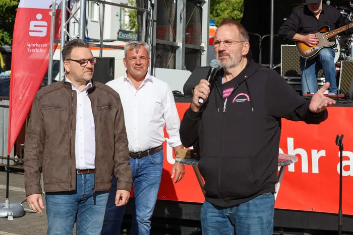 Ein Mann spricht mit einem Mikrofon auf einer Bühne beim Brückenfest, während zwei Männer neben ihm stehen.