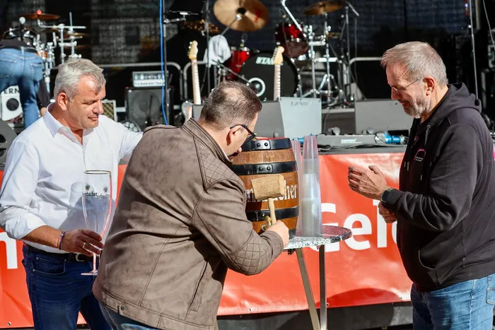 Bürgermeister Michael Joithe sticht mit einem Holzhammer ein kleines Holzfass an. Zwei Männer beobachten ihn vor einer Bühne mit Musikinstrumenten.