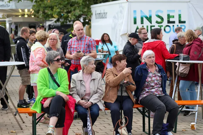 Menschen sitzen auf einer orangefarbenen Bank, im Hintergrund stehen weitere Besucher vor einem weißen Transporter mit der Aufschrift „INSEL TOUR“.
