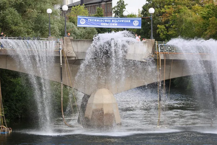 Wasserfontänen sprühen von einer Betonbrücke in einen Fluss, an der ein blaues Banner mit der Aufschrift „TECHNISCHES HILFSWERK ISERLOHN“ hängt.