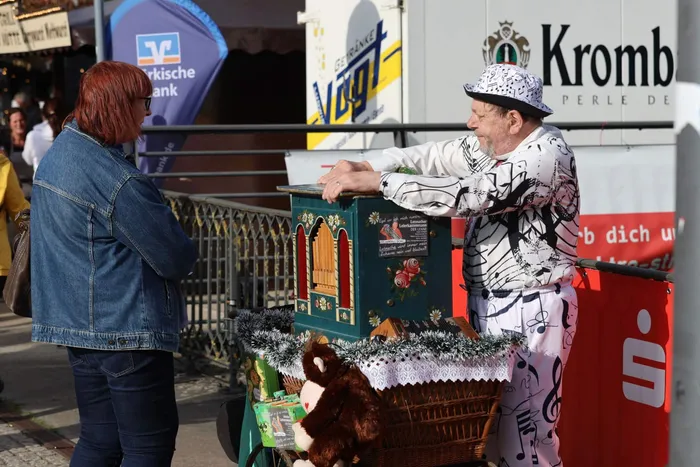 Ein Mann in einem weiß-schwarzen Outfit mit Musiknoten bedient eine kleine, verzierte Orgel, während eine Frau ihm zuschaut.