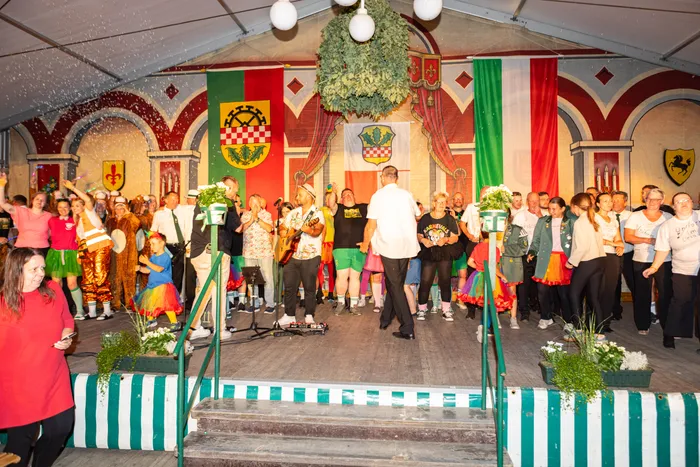 Menschen in bunten Kostümen und Alltagskleidung feiern auf einer Bühne bei einem Schützenfest vor Wappen und Flaggen.