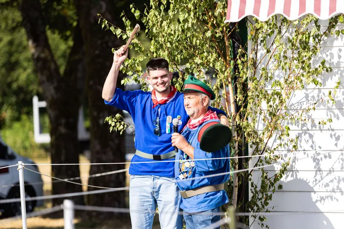 Zwei Männer in blau-roter bzw. blau-grauer Tracht stehen lächelnd vor einem festlich geschmückten Stand. Der jüngere hebt die Hand, während der ältere in ein Handmikrofon spricht.