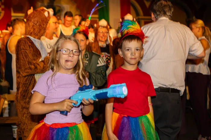 Zwei Kinder in bunten Tutus stehen im Vordergrund, ein Mädchen hält eine blaue Spielzeugpistole. Im Hintergrund sind weitere Personen in Kostümen zu sehen.