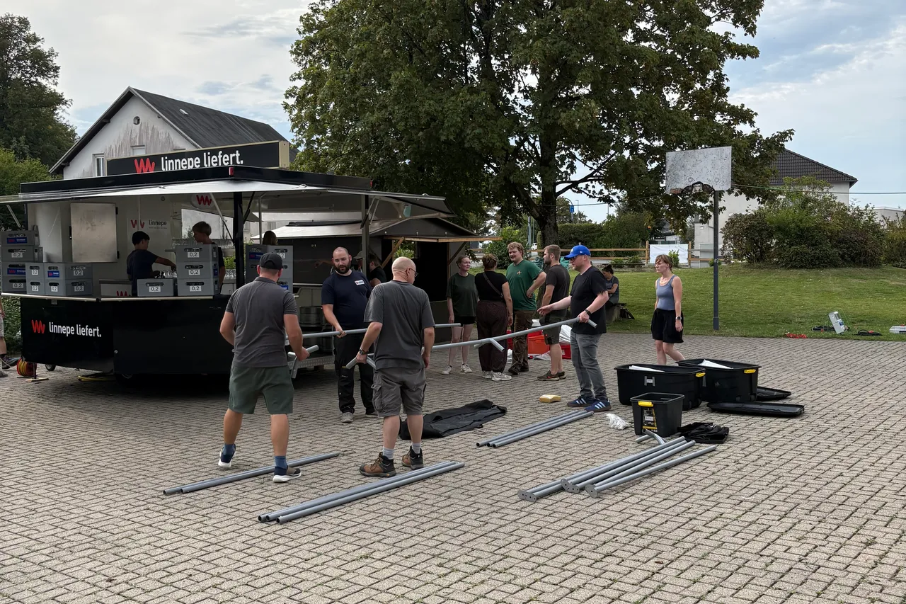 Helfer bereiten ein Sommerfest vor und hantieren mit Metallstangen auf einem gepflasterten Platz vor einem schwarzen Imbisswagen.