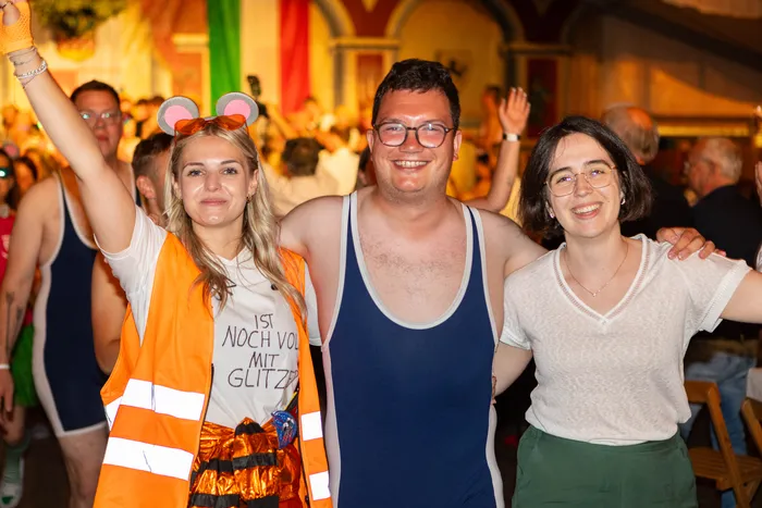 Drei lächelnde Personen posieren bei einer Feier der Hülscheider Schützen, eine Frau trägt eine orangefarbene Warnweste und Mäuseohren.