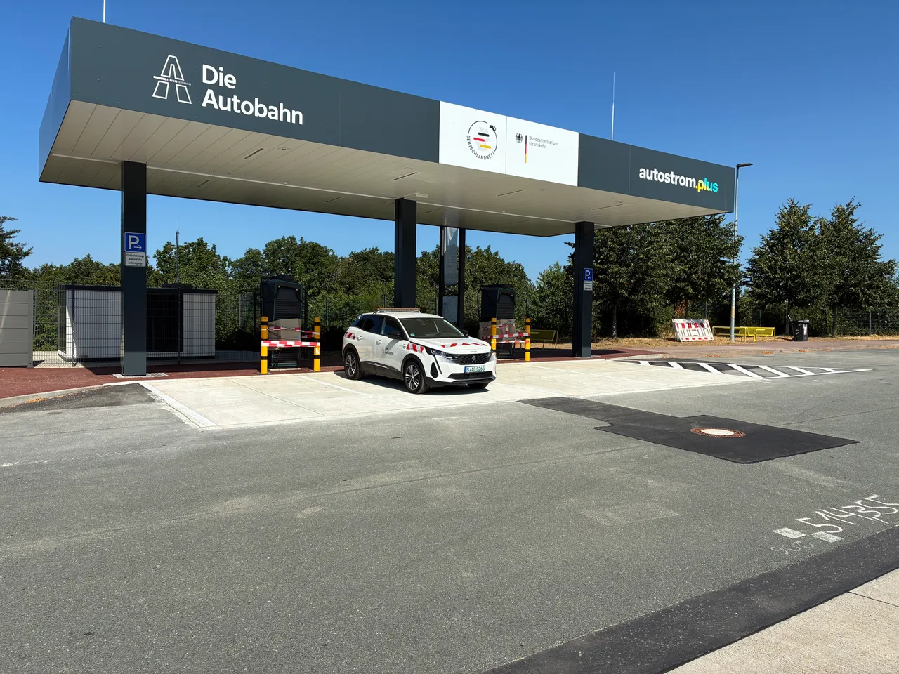 Ein weißes Auto an einer Schnellladestation unter einem großen Überdach mit Logos der Autobahn GmbH und autostrom.plus.