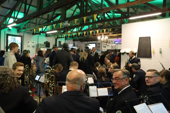 Eine Menschenmenge in einem hellen Raum mit Holzbalken lauscht einem Musikkorps in Uniformen. Im Hintergrund eine Tafel mit Schrift und Porträts.