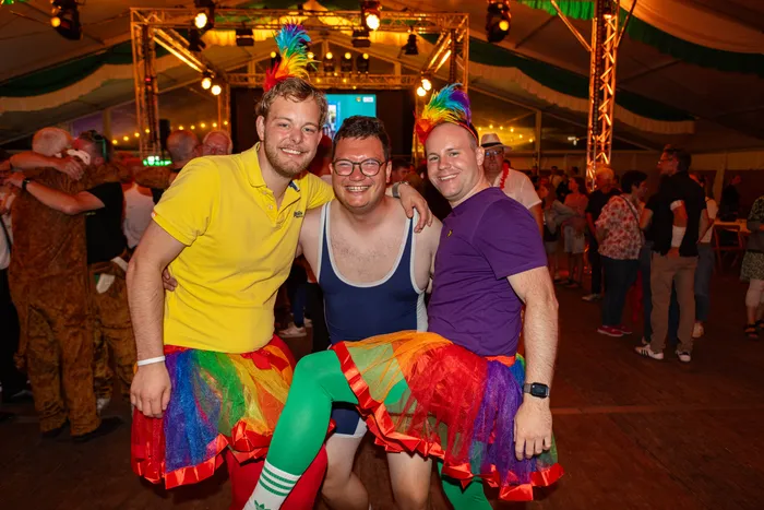 Drei lächelnde Männer in bunten T-Shirts und Regenbogen-Tutus posieren in einem festlich beleuchteten Festzelt.