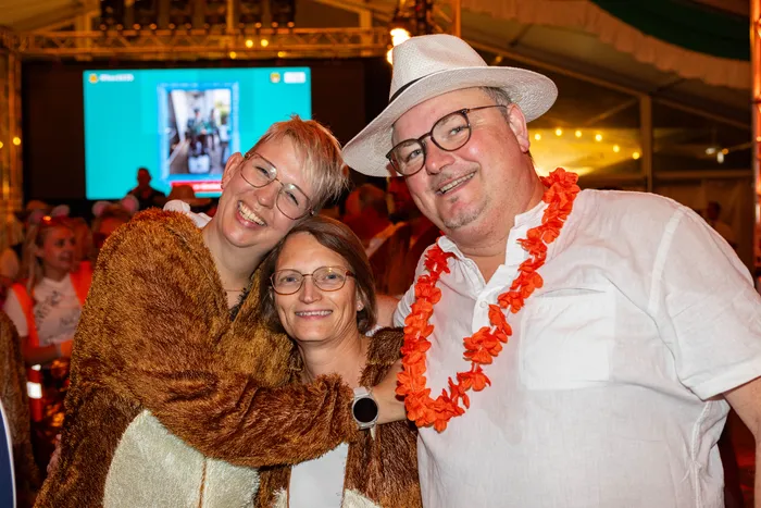Drei lächelnde Personen posieren bei einem Schützenfest, eine in einem braunen Fellkostüm und eine mit weißem Hut und orangefarbener Blumenkette.