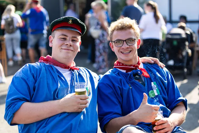Zwei junge Männer in blauem Hemd und mit roten Halstüchern lächeln in die Kamera, einer trägt eine Schützenmütze und hält ein Bierglas.