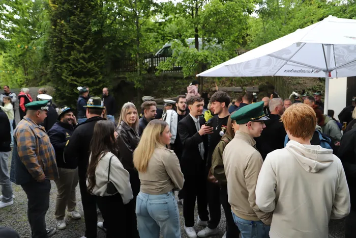 Eine Gruppe von Menschen steht im Freien unter einem weißen Zelt zusammen, einige tragen Uniformen mit grünen Mützen. Im Hintergrund sind Bäume und ein grauer PKW zu sehen.