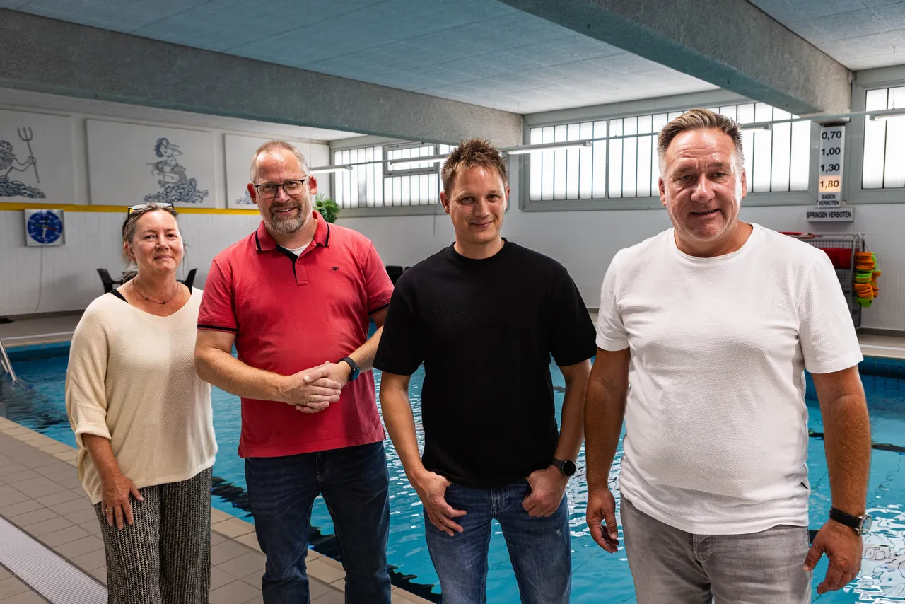 Vier Personen stehen lächelnd am Rand eines Schwimmbeckens in einer Schwimmhalle.