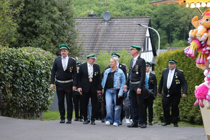 Eine blonde Frau in Jeansjacke geht lächelnd umgeben von Männern in dunklen Anzügen und grünen Schützenhüten auf einer Straße. Im Hintergrund sind Häuser und ein Jahrmarktstand mit Stofftieren zu sehen.