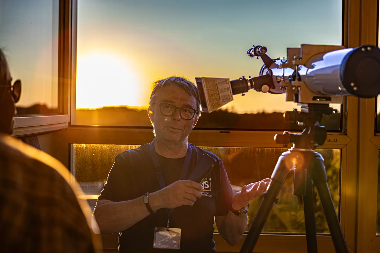 Ein Mann erklärt vor einem Fenster bei Sonnenuntergang ein Teleskop auf einem Stativ.