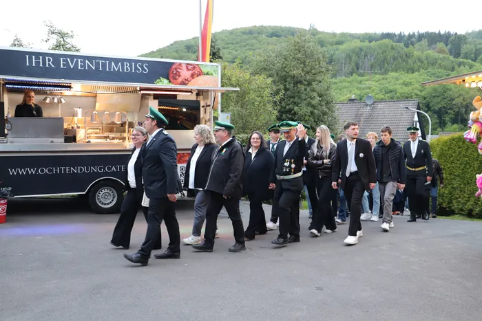 Eine Gruppe von Menschen in festlicher Kleidung geht an einem Imbisswagen vorbei, auf dem „Ihr Eventimbiss“ steht. Im Hintergrund eine Hügellandschaft.